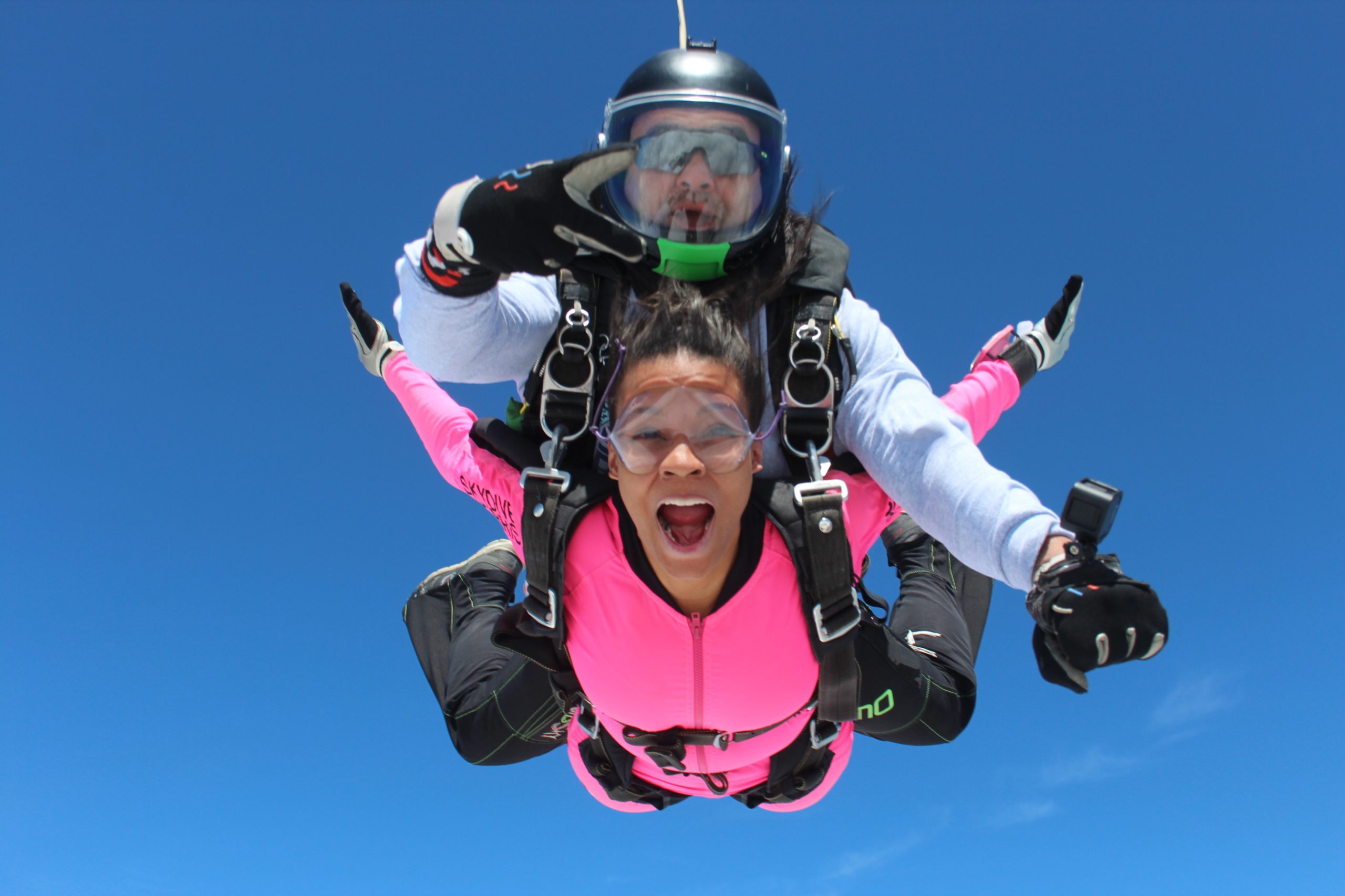 A girl in a pink jumpsuit smiles during free fall on a tandem skydive.