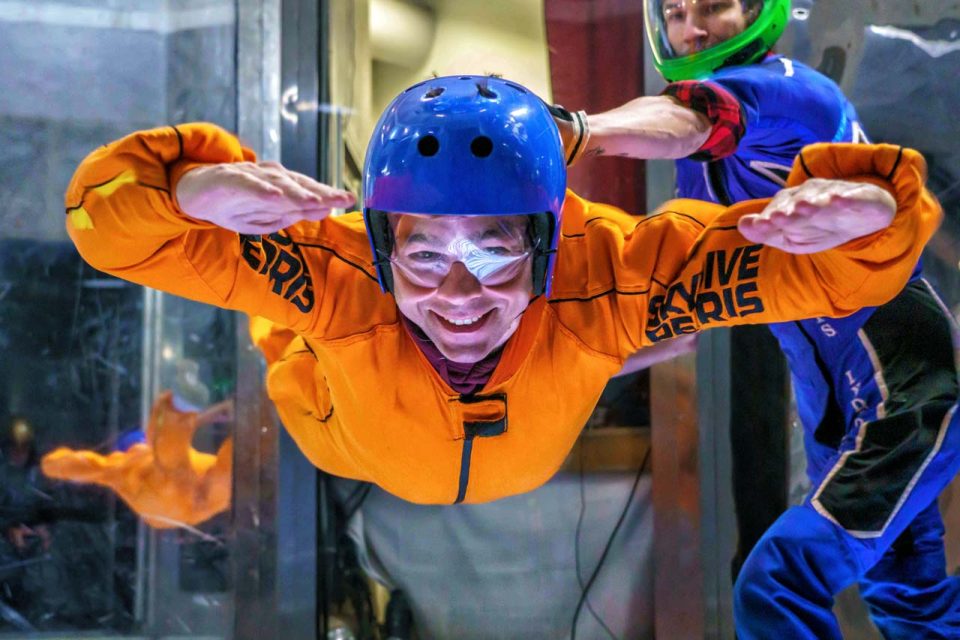 an adult man in an orange jumpsuit smiles while flying in the tunnel.